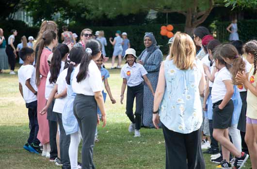children and teachers stand in a green space clapping, smiling, singing and enjoying outdoor activities in the sun