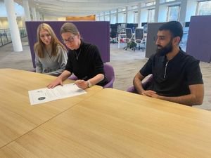 A woman sits at a table signing a document. A young man and a young woman are watching her.