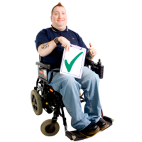 A man sitting in a wheelchair with his thumb up and holding a sign with a big green tick