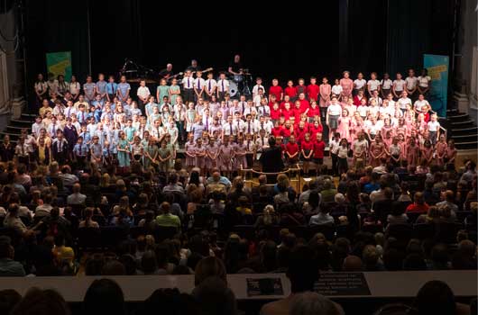 Image of the Dorking Halls theatre showing many rows of school children singing alongside a live band and the conductor performing to a large seated audience.