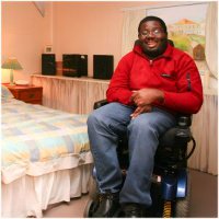 Man in a power wheelchair smiling in a tidy bedroom with a large bed and bright decor.
